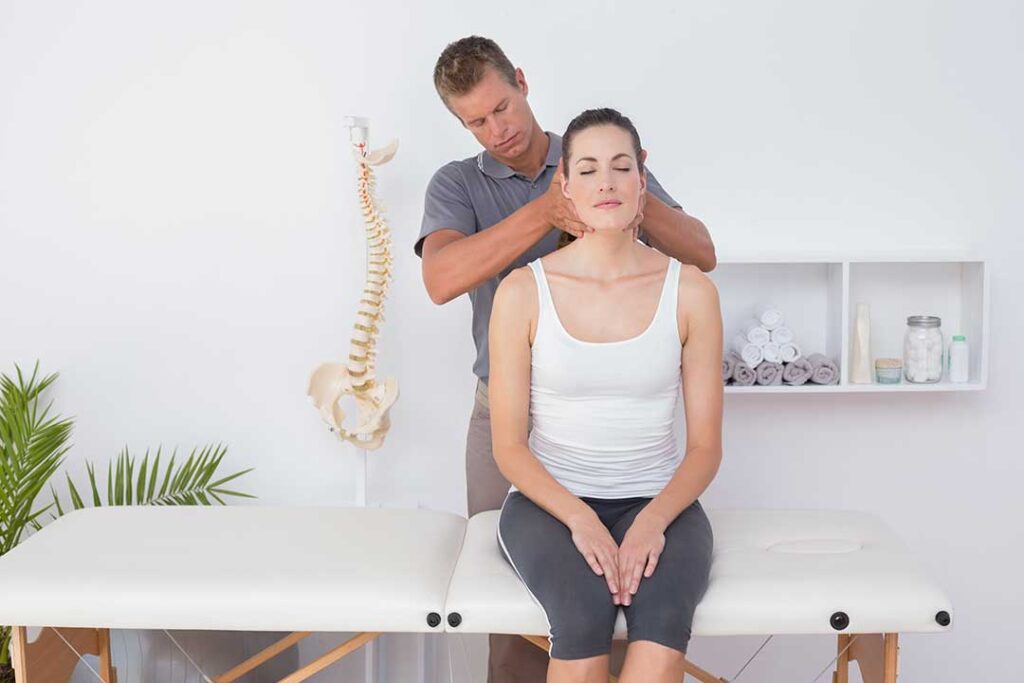 A chiropractor gently assessing a patient's neck while the patient sits upright in a clinical setting.